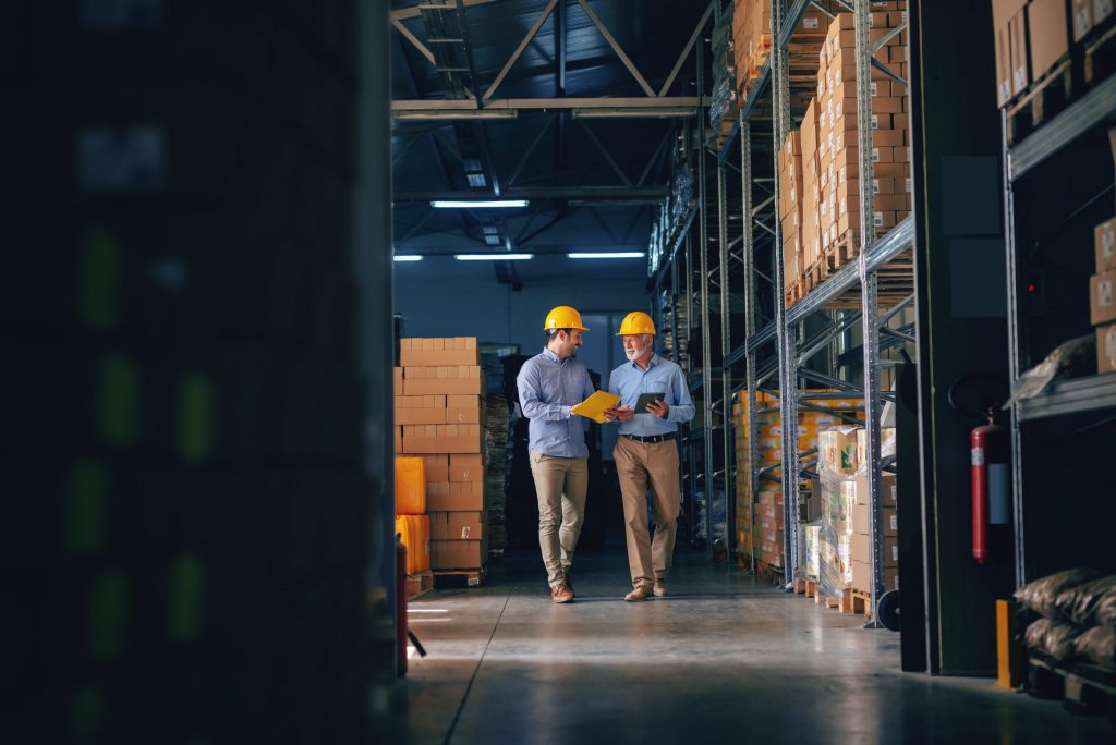 Two men in hard hats walk through shelving in a manufacturing facility. 
