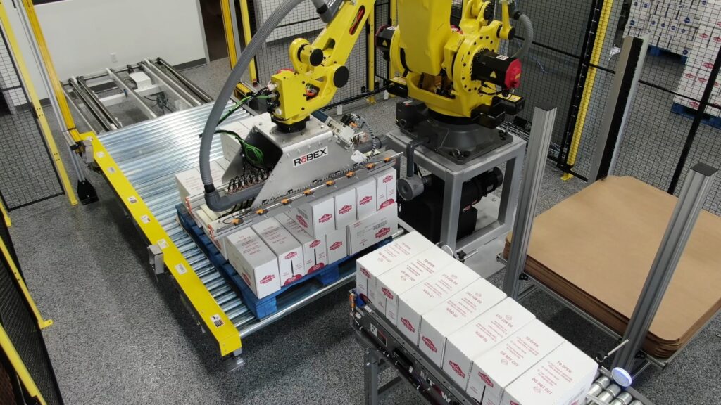 A yellow robot moves cardboard boxes into a stack on a pallet