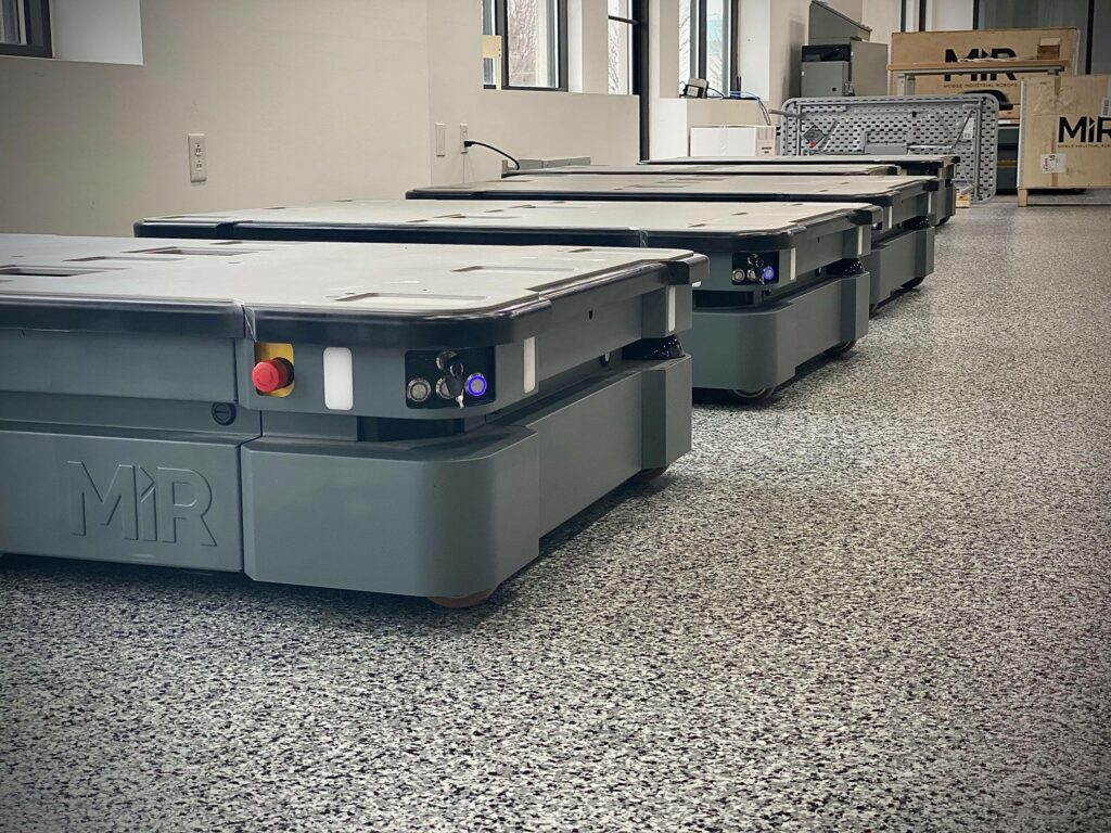 Five mobile industrial robots sit on a gray epoxy floor.