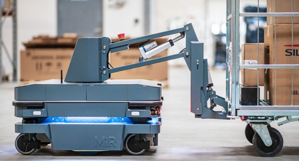 A gray MiR robot pulls a stainless steel cart on a white floor.