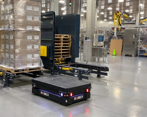 A black autonomous mobile robot prepares to lift a pallet full of boxed items for transport on a concrete floor.